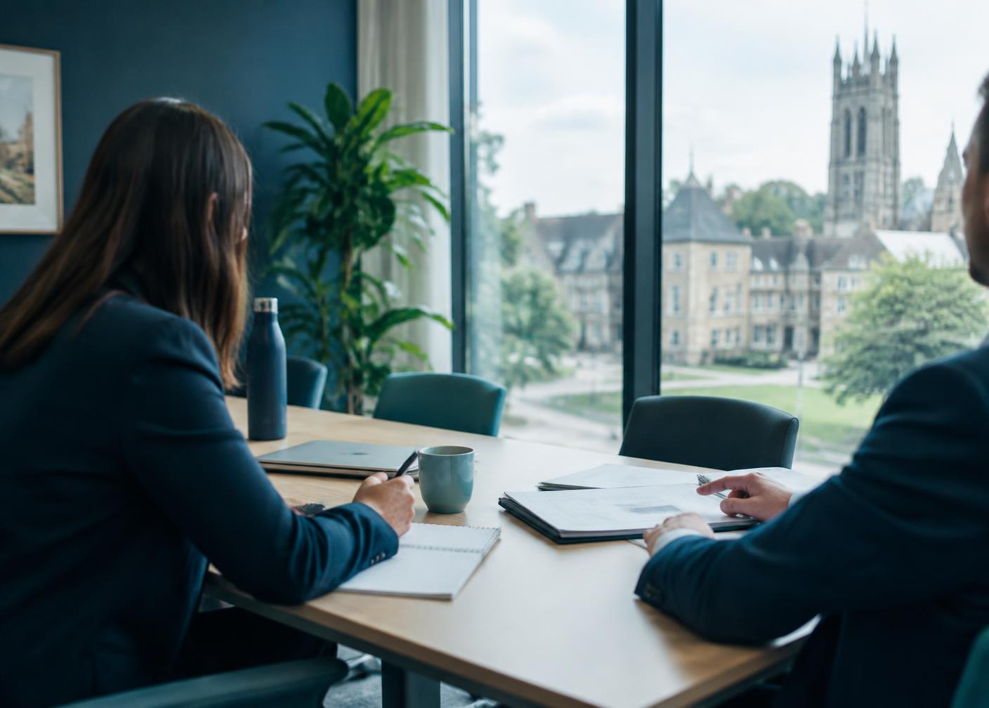 Two professionals in discussion in a meeting room overlooking a UK campus